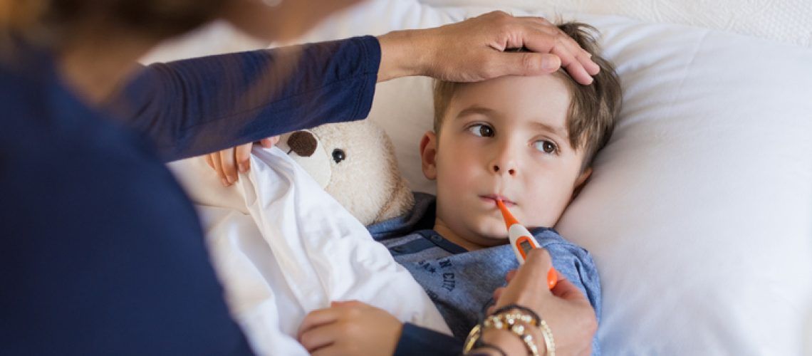 Sick boy with thermometer laying in bed and mother hand taking temperature. Mother checking temperature of her sick son who has thermometer in his mouth. Sick child with fever and illness while resting in bed.